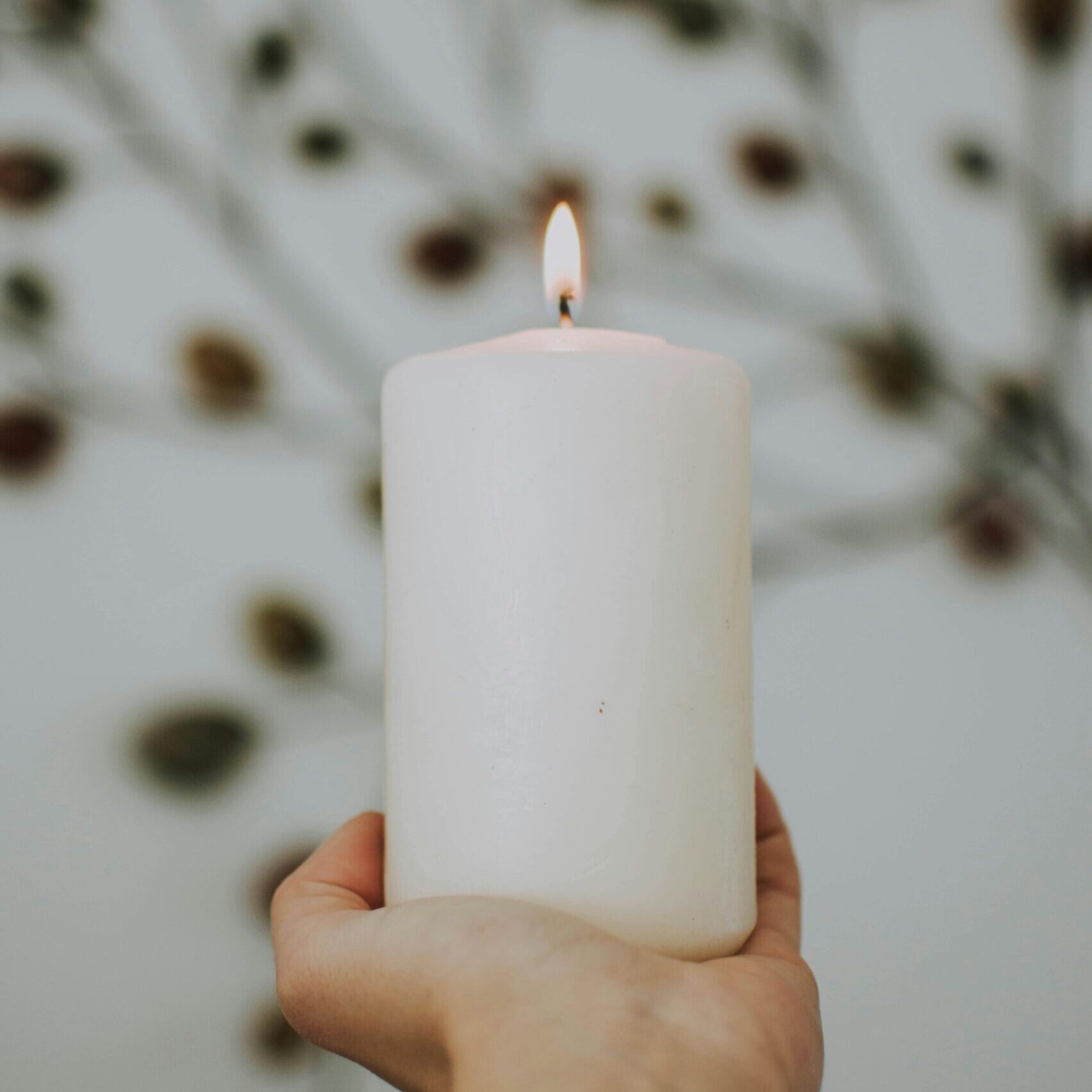 A hand holds a lit white candle against a blurred, artistic backdrop, creating a peaceful ambiance.